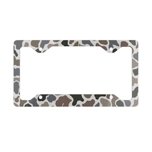 May include: License plate frame with a camouflage pattern in shades of gray, brown, and white. The frame has a rectangular shape with rounded corners and mounting holes.