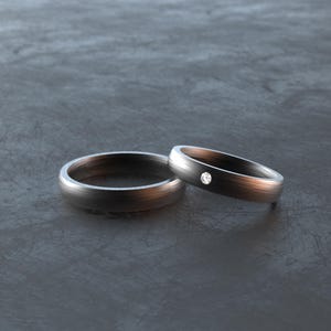 Könnte beinhalten: Zwei dunkelgraue Eheringe. Ein Ring hat einen kleinen, runden Diamanten. Die Ringe haben eine gebürstete Metalloberfläche und sind vor einem dunkelgrauen Hintergrund platziert. Die Ringe sind wahrscheinlich für eine Hochzeit oder einen Verlobung.