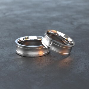 Könnte beinhalten: Zwei silberne Ringe mit gebürsteter Metalloberfläche. Ein Ring hat drei kleine Diamanten. Die Ringe sind vor einem dunkelgrauen Hintergrund platziert, wobei sich das Licht auf den Metalloberflächen spiegelt.