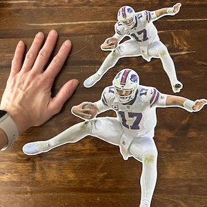 May include: Two cardboard cutouts of an American football player wearing a white jersey with blue and red accents. The player is wearing a helmet and is in a throwing motion. The cutouts are on a wooden surface.