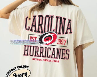 Puede incluir: Una camiseta blanca con un logotipo de los Carolina Hurricanes y el texto "Carolina Est 1997 Hurricanes National Hockey League" impreso en la parte delantera. La camiseta es de color marfil.