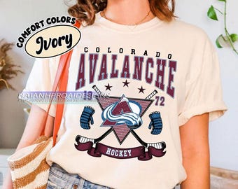 May include: A white t-shirt with a burgundy and blue Colorado Avalanche hockey logo. The logo features a stylized 'A' with a wave design, two crossed hockey sticks, and the text 'Colorado Avalanche' and 'Hockey'.