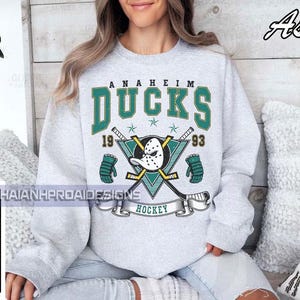 Op de afbeelding: Een lichtgrijs sweatshirt met het Anaheim Ducks-logo in teal en wit. Het ontwerp bevat de teamnaam, een hockeymasker, gekruiste hockeysticks en de jaren 1993 en 1993. Het woord "HOCKEY" is op een banner gedrukt.