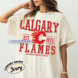 May include: Ivory t-shirt with the words "CALGARY FLAMES" in red, and a flaming "C" logo. The shirt also has "EST. 1972" and "NATIONAL HOCKEY LEAGUE". The shirt is from Comfort Colors.