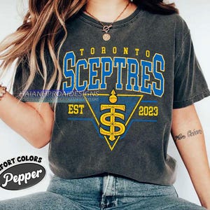 以下が含まれることがあります： 青と黄色のグラフィックデザインが施された濃いグレーのTシャツ。グラフィックには「Toronto Sceptres」というテキストと、「EST 2023」というテキストが書かれたスタイリッシュなロゴが描かれています。