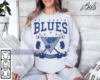 May include: A light gray sweatshirt featuring the text "ST. LOUIS BLUES" in blue and gold, along with hockey-themed graphics. The design includes crossed hockey sticks, a logo, and the year "1967". The word "HOCKEY" is printed on a blue banner.