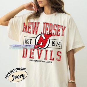 Może przedstawiać: Biały T-shirt z czerwono-czarnym logo New Jersey Devils i tekstem "NEW JERSEY EST. 1974 DEVILS NATIONAL HOCKEY LEAGUE". Koszulka ma etykietę "COMFORT COLORS Ivory".