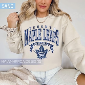 Può includere: Felpa color sabbia con la scritta blu "TORONTO MAPLE LEAFS ESTABLISHED 1917". Il design include una grafica a foglia d'acero e il marchio "HAIANHPROAIDESIGNS".