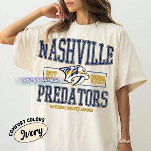 May include: A white t-shirt with a vintage-style graphic of the Nashville Predators hockey team. The graphic features the team's logo, the words "Nashville Predators", "Est. 1998", and "National Hockey League". The shirt has a distressed look and is printed with the words "Comfort Colors" and "Ivory" in a circle on the bottom left.