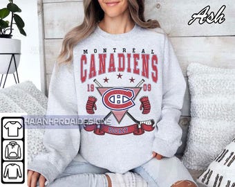 Puede incluir: Una sudadera gris claro con la inscripción "MONTREAL CANADIENS" en rojo, y un gráfico de hockey. El gráfico incluye palos de hockey cruzados, un logotipo y los años "1909". La palabra "HOCKEY" está en una pancarta roja.