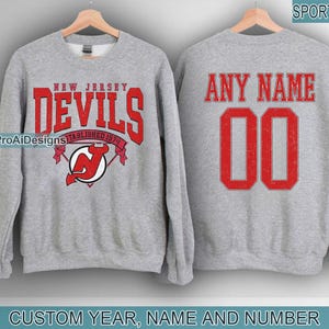 May include: A gray sweatshirt with the text "NEW JERSEY DEVILS" in red, along with a team logo. The back of the sweatshirt displays "ANY NAME" and the number "00" in red. The words "CUSTOM YEAR, NAME AND NUMBER" are at the bottom.