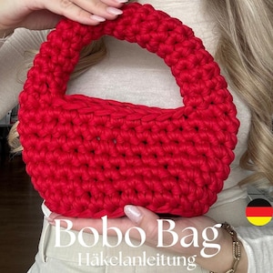 Peut inclure: Un sac en crochet rouge vif avec une anse. Le sac est tenu par la main d'une personne. Le texte "Bobo Bag Häkelanleitung" est visible sous le sac.