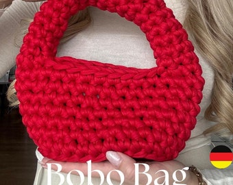 May include: A bright red crocheted bag with a handle. The bag is held by a person's hand. The text "Bobo Bag Häkelanleitung" is visible below the bag.