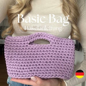 Könnte beinhalten: Eine lilafarbene Häkel-Tasche mit Henkel. Die Tasche wird von einer Person gehalten, die einen hellbraunen Pullover und eine blaue Jeans trägt. Der Text "Basic Bag Häkelanleitung" ist auf dem Pullover zu sehen.