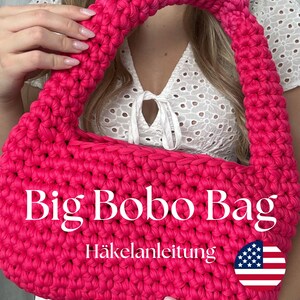 Könnte beinhalten: Eine leuchtend pinkfarbene Häkeltasche mit einem großen Henkel. Die Tasche trägt den Text "Big Bobo Bag" und "Häkelanleitung". Die Tasche ist mit einer kleinen amerikanischen Flagge verziert.