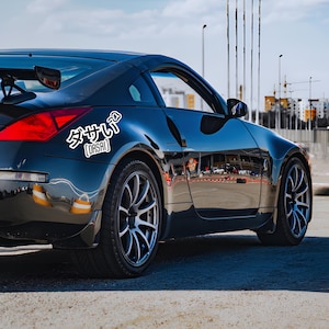Puede incluir: Un Nissan 350Z negro con una pegatina blanca que dice "ダサい [DASAI]" en la parte trasera del coche. El coche está aparcado en una superficie pavimentada con un fondo borroso.