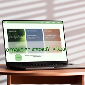 以下が含まれることがあります： 「to make an impact?」と「Contact us」のテキストを含む、緑と白の要素を持つウェブサイトを表示するラップトップ。画面には、炭素削減、ブランド変革、ESGフレームワークに関する情報が表示されています。ラップトップは、丸い茶色のテーブルの上にあります。