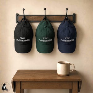 May include: Three baseball caps in black, green, and navy blue hang on a wooden rack. Each cap has the text "Over Caffeinated" in white. A mug of coffee sits on a small wooden table below the hats.