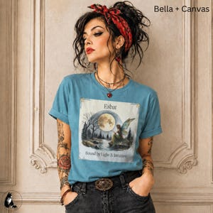 Może przedstawiać: Niebieski t-shirt z nadrukiem graficznym przedstawiającym leśną scenę w świetle księżyca z wróżką, napisem Esbat oraz Bound by Light & Intuition. Koszulka jest zestawiona z czerwoną wzorzystą chustą na głowę, wielowarstwowym naszyjnikiem i ciemnymi dżinsami.