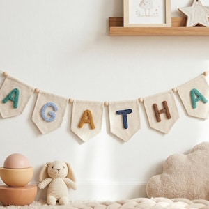Puede incluir: Una guirnalda con el nombre "AGATHA" con letras coloridas y texturizadas cuelga de una pared blanca. Debajo, un juego de cuencos de madera, un conejito de ganchillo y una almohada en forma de nube crean un ambiente de guarder&iacute;a.
