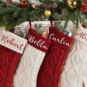 Peut inclure: Quatre bas de Noël personnalisés sont suspendus à une cheminée décorée de branches de sapin et d'ornements. Les bas sont en tricot torsadé rouge et blanc avec des noms brodés sur les revers : Robert, Bella, Carlin et Alison.