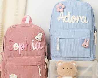 May include: Two corduroy backpacks, one pink and one light blue, with personalised names. The pink backpack has the name "Sophia" and the blue backpack has the name "Adora". Both have decorative appliques and a front pocket.