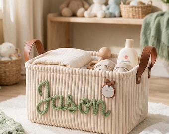 Puede incluir: Una cesta de almacenamiento rectangular de pana color crema con asas de piel sintética marrón. El nombre Jason está escrito en letra cursiva de hilo verde en la parte frontal, junto a un pequeño parche de cara de oso. Contiene una tela blanca.