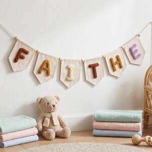 Puede incluir: Un cartel con la palabra "FAITHE" cuelga sobre un oso de peluche, mantas y un juguete de madera. El cartel tiene banderines beige con letras coloridas y texturizadas. La escena est&aacute; ambientada en una pared blanca, creando un ambiente acogedor.