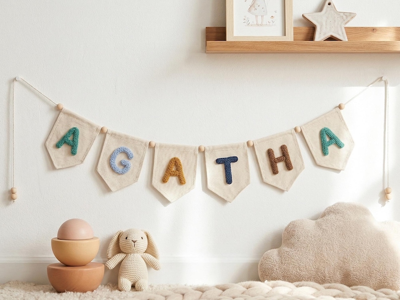 Puede incluir: Una guirnalda con el nombre "AGATHA" con letras coloridas y texturizadas cuelga de una pared blanca. Debajo, un juego de cuencos de madera, un conejito de ganchillo y una almohada en forma de nube crean un ambiente de guarder&iacute;a.