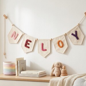 Puede incluir: Un cartel con el nombre "NELOY" cuelga de una pared. Las letras son de color rosa, burdeos y naranja sobre banderines blanquecinos. Un conejito de peluche, anillos de madera apilables y libros est&aacute;n sobre una superficie de madera.