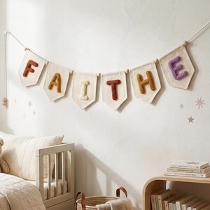 Puede incluir: Un cartel con la palabra "FAITHE" cuelga de una pared. Cada letra es de un color diferente, con un fondo color crema. El cartel est&aacute; ensartado en un cord&oacute;n blanco con cuentas de madera. La habitaci&oacute;n tiene una paleta de colores neutros.
