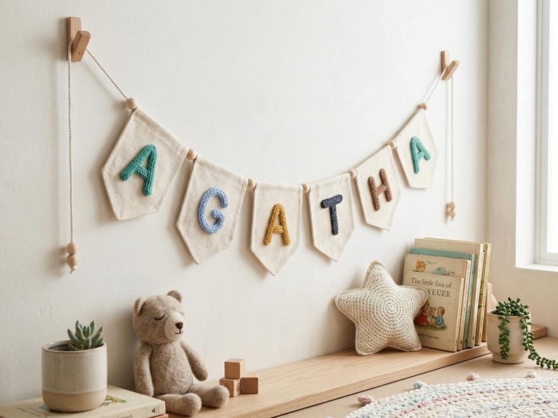Puede incluir: Un cartel con banderas de tela individuales que deletrean "AGATHA", cada bandera muestra una letra en un color diferente. Las letras son en turquesa, azul, amarillo, marr&oacute;n y azul marino. El cartel est&aacute; colgado en una pared blanca sobre una estanter&iacute;a de madera.