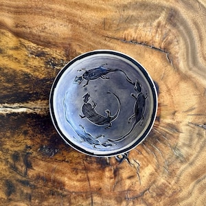 May include: A circular ceramic bowl with a light grey interior featuring a dark black rim and a hand-drawn illustration of four black lizards arranged in a circular pattern around the centre of the basin, showcasing detailed scale textures.