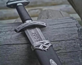 May include: A detailed image of a Viking sword with a dark wooden handle and a silver-colored blade. The blade has etched patterns and the word "NOR" is visible. The sword is in a dark wooden scabbard with silver accents. The sword is resting on a wooden surface.