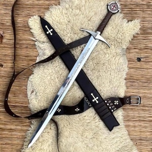 Könnte beinhalten: Ein mittelalterliches Schwert mit silberner Klinge und braunem Griff, das auf einer schwarzen Scheide und einem braunen Ledergürtel liegt. Die Scheide und der Gürtel sind mit Kreuzsymbolen verziert. Der Knauf des Schwertes hat ein rundes Emblem.