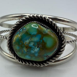 May include: A silver cuff bracelet featuring a large, polished, oval-shaped turquoise stone with blue and green marbling. The stone is set in a twisted rope-style silver bezel, and the band consists of two smooth silver wires and one twisted wire.
