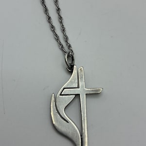 May include: A silver-toned metal pendant necklace featuring a cross symbol integrated with a stylized flame design. The pendant hangs from a twisted rope chain, showcasing a smooth, polished finish on the metallic surface of the cross and flame.