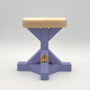 May include: A handstand cane base with a light-coloured wooden top and a lavender-coloured X-shaped support structure. A small circular wooden plaque is attached to the centre of the support, featuring the text VertiBase Fitness Equipment.