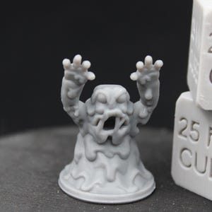 May include: A gray, stylized monster figurine with arms raised and an open mouth. The figurine is next to two stacked white cubes with the text "25 MM CUBE" on the sides. The figurine has a melted appearance.
