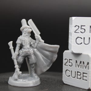 May include: A gray resin miniature figure of a character wearing a tall hat, goggles, and a cape, holding a long staff and a serrated blade. Next to the figure are two stacked white cubes, each clearly marked with the text 25 MM CUBE.