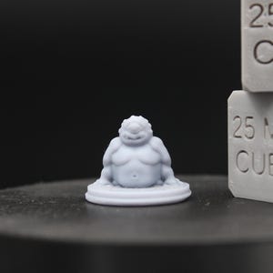 May include: A light gray, unpainted resin miniature of a stout, single-eyed creature with a round belly, seated on a circular base. Next to the figure are two stacked gray blocks, each clearly embossed with the text 25 MM CUBE.