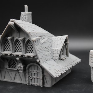 May include: A gray 3D printed miniature building with a shingled roof, arched lattice windows, and a wooden door, positioned next to two stacked 25 mm cube blocks for scale. The structure features detailed timber framing and a chimney.