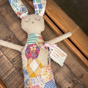 May include: A handcrafted patchwork bunny doll with closed eyes and outstretched arms. The bunny's ears feature floral and geometric patterns. The body is a patchwork of various fabrics in pinks, blues, yellows, and greens. A tag reads "Country Girl Corner Limited Edition Patchwork Bunny."