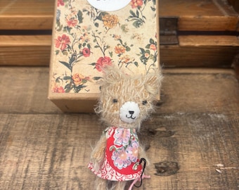 May include: A small, fuzzy, light brown teddy bear wearing a red dress with a pink and white floral pattern. The bear has a white muzzle with a stitched nose and mouth. A white tag attached with a pink string reads "Sally" in brown letters.