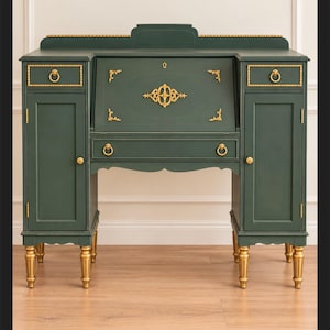 May include: A dark green secretary desk featuring a drop-down writing surface, two side cabinets, and two small upper drawers. The piece is accented with gold-toned metal hardware, including ring pulls, decorative filigree, and four tapered legs.
