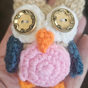 May include: A handmade crochet owl with a tan head, white face, and pink body. The owl has gold button eyes and an orange beak and feet. The wings are blue. This whimsical owl is a unique handmade item.
