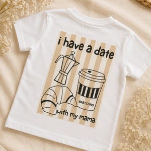 May include: A white short-sleeved t-shirt featuring a rectangular graphic with beige vertical stripes. The design includes black line art of a moka pot, a croissant, and a coffee cup labelled "BABYCCINO" with the text "i have a date with my mama".