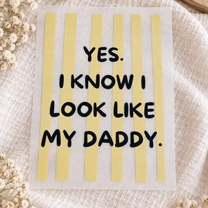 Op de afbeelding: Een rechthoekig wit transfervel met verticale lichtgele strepen en de zwarte tekst "YES. I KNOW I LOOK LIKE MY DADDY." De letters zijn in een vet, afgerond schreefloos lettertype en zijn gecentreerd over het gestreepte achtergrondpatroon.