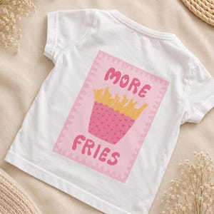 Puede incluir: Una camiseta blanca de manga corta con un gráfico rectangular rosa en la parte delantera. El gráfico muestra un recipiente rosa lleno de patatas fritas amarillas y el texto MORE FRIES en letras rosas. El recipiente tiene un patrón de corazones.