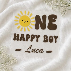 Könnte beinhalten: Ein quadratisches Schild aus klarem Acryl mit einer gelben Sonne mit lächelndem Gesicht, die den Buchstaben O im Wort ONE ersetzt. Darunter steht der Text HAPPY BOY und der Name Luca in dunkelbrauner Schrift mit zwei Linien.